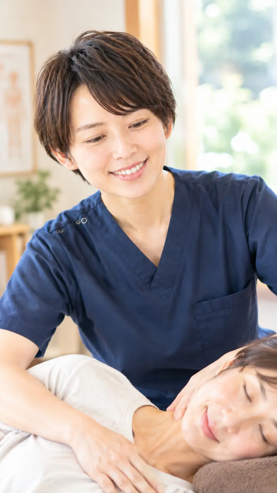明るい院内で、紺色のユニフォームの女性スタッフが微笑みながら女性患者の首まわりを施術している様子